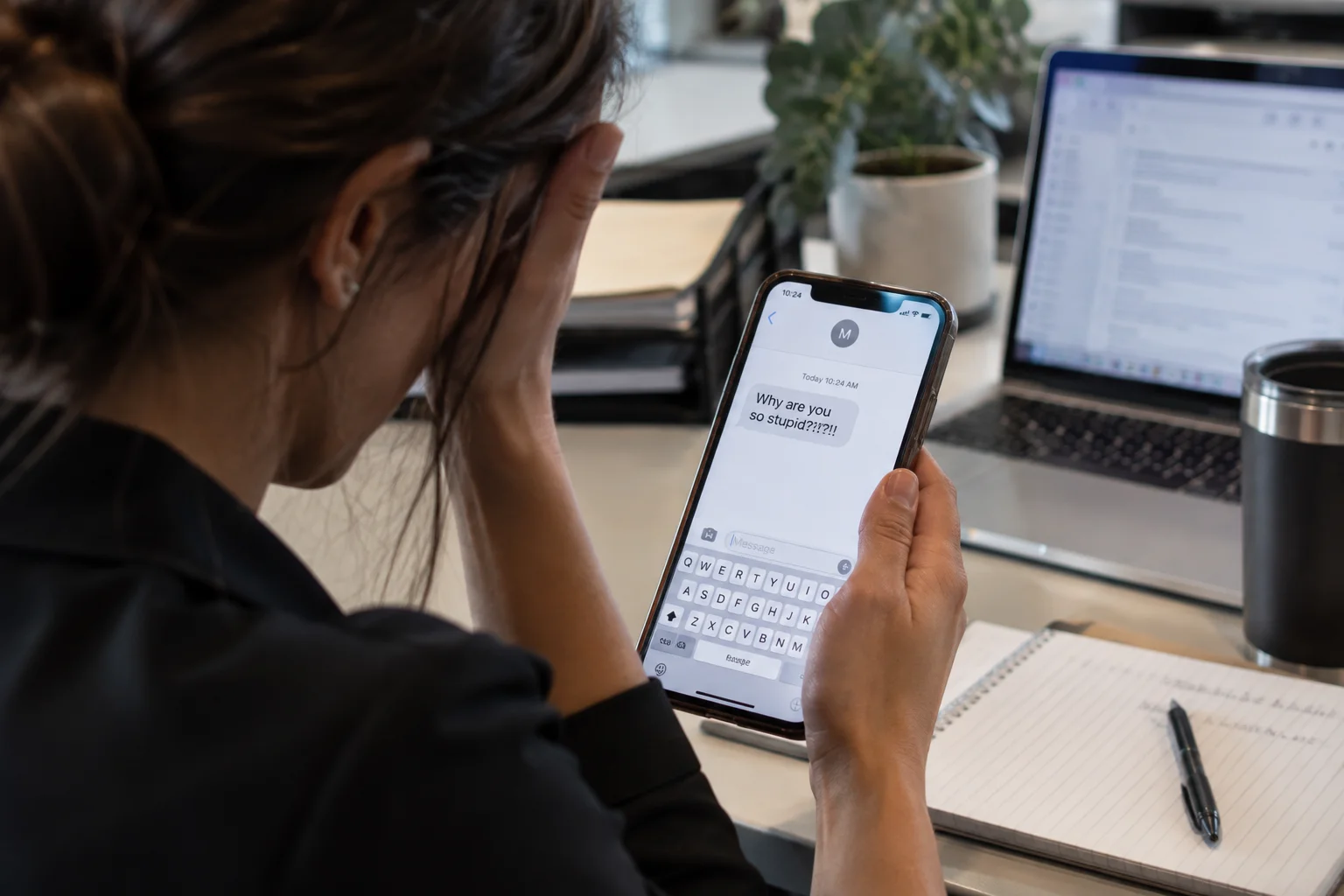 Distressed employee at her desk reading a hostile text message on her phone